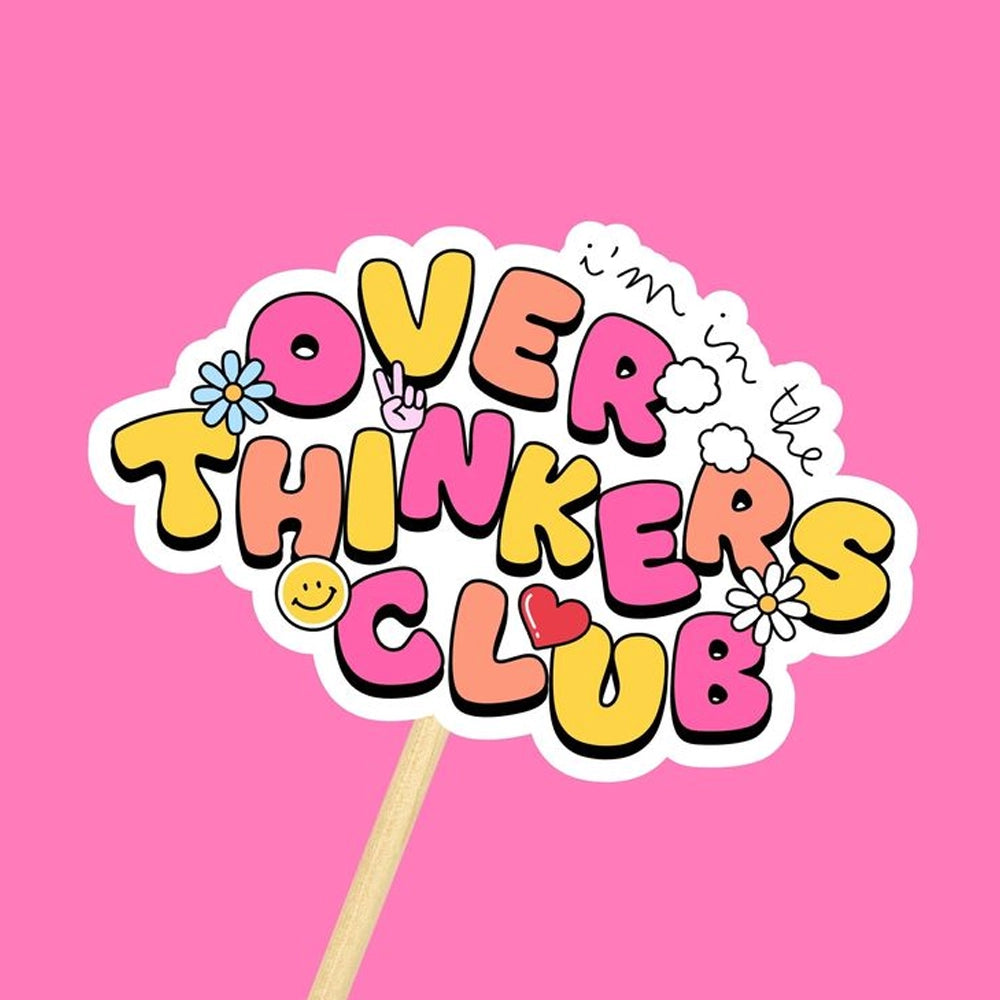 Colorful hand-drawn sign reading ‘Over Thinkers Club’ in playful bubble letters, decorated with flowers, a peace sign, smiley face, heart, and doodles, on a stick against a bright pink background.