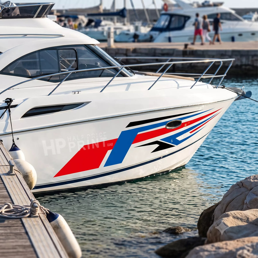 A white motor yacht docked at a harbor, featuring a bold red, blue, and black boat graphic stripe design on its side. The boat is pictured in a real-world marina environment with water, rocks, and other boats in the background.