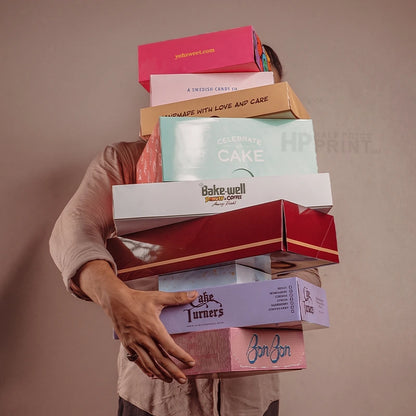 A person in a beige shirt carries a tall stack of colorful bakery cardboard gift boxes, including boxes for Cake Warehouse, Bake Well, and Jon Jon, against a solid taupe background.