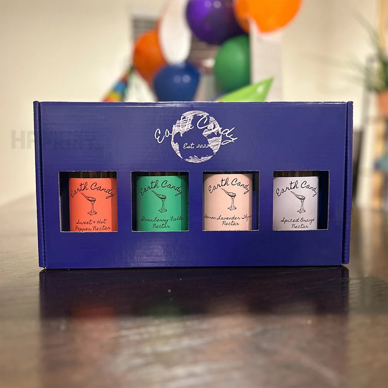 A blue gift box from Earth Candy containing four small colorful bottles of house blends, displayed on a dark wooden table with a blurred festive background of balloons.