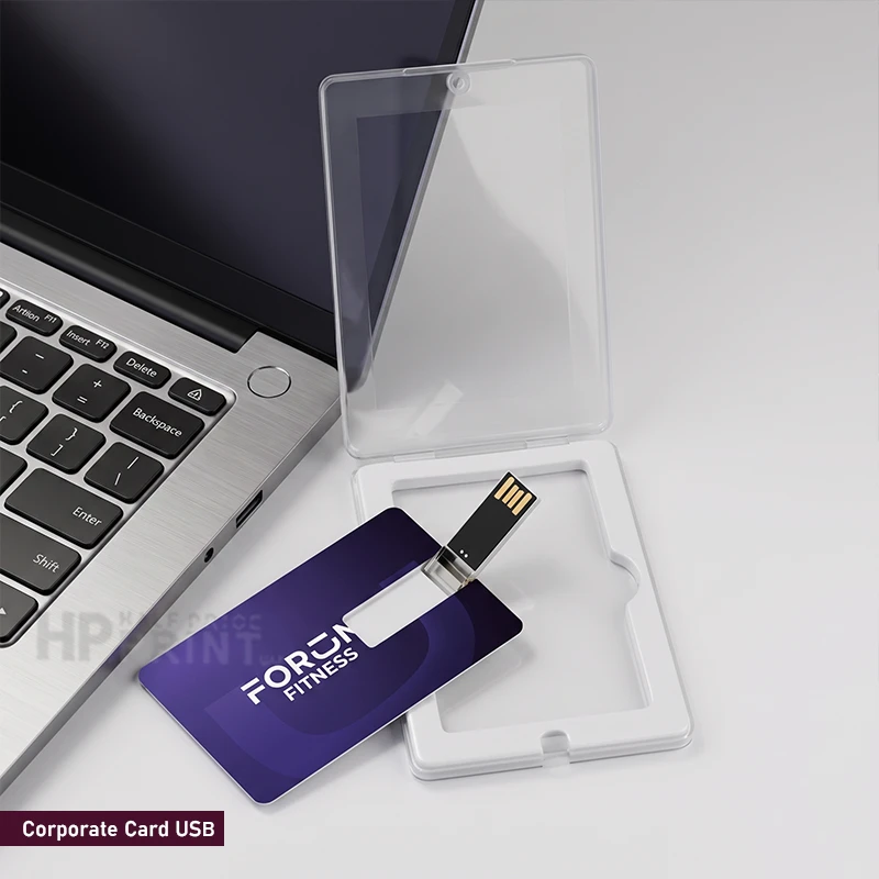 A purple corporate card USB drive with a metal USB connector, shown next to its clear plastic protective case and white storage tray on a light gray surface, with part of a laptop visible in the background.