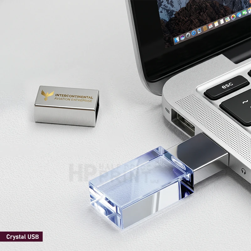 A silver and clear acrylic USB flash drive shown plugged into a silver laptop on a light gray surface, with the matching cap placed nearby. The cap is printed with the Intercontinental Aviation Enterprise logo and name.