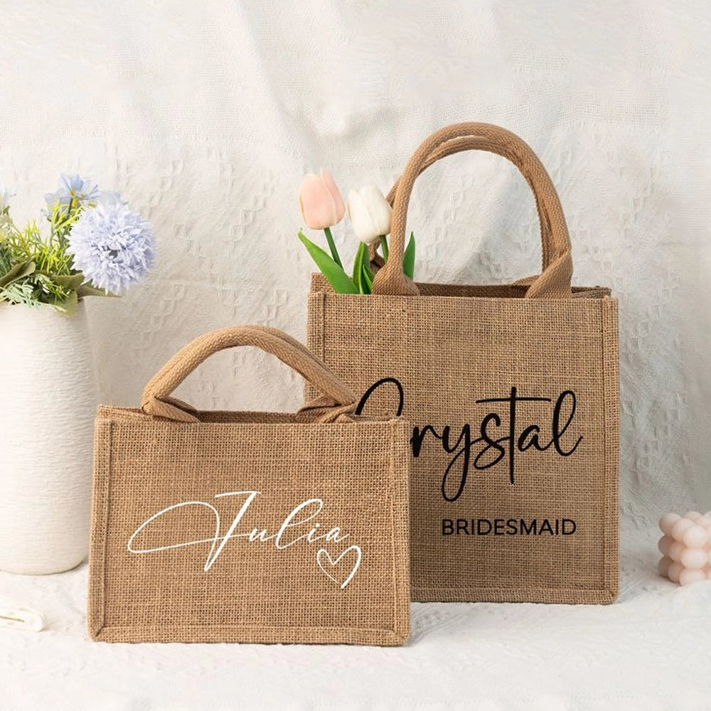 Two small burlap tote bags styled with flowers, customized with names and wedding roles in script lettering, shown on a light textured background for a bridesmaid gift display.