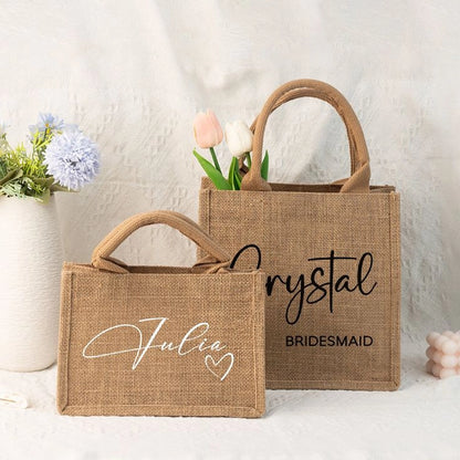 Two small burlap tote bags styled with flowers, customized with names and wedding roles in script lettering, shown on a light textured background for a bridesmaid gift display.
