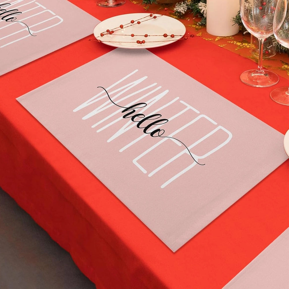 Holiday table setting with a red tablecloth, featuring a blush-colored placemat that reads ‘hello’ in black script. A white plate with a red beaded garland sits above the placemat, alongside wine glasses and festive greenery.