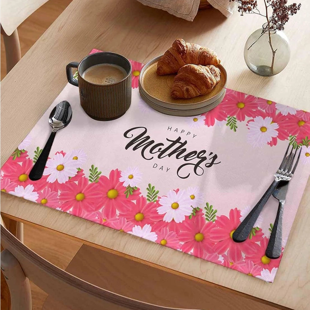 Pink floral placemat on a dining table with the text “Happy Mother’s Day,” set with a coffee mug, croissants on a plate, cutlery, and a small vase.
