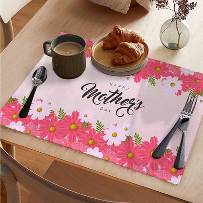 Pink floral placemat on a dining table with the text “Happy Mother’s Day,” set with a coffee mug, croissants on a plate, cutlery, and a small vase.