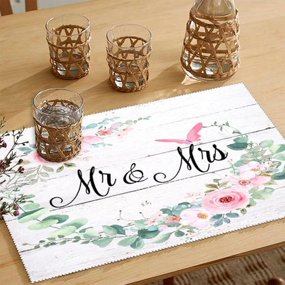 Rustic floral table mat on a light wood table with the text “Mr & Mrs,” surrounded by greenery and pink flowers, with glass tumblers wrapped in wicker holders.