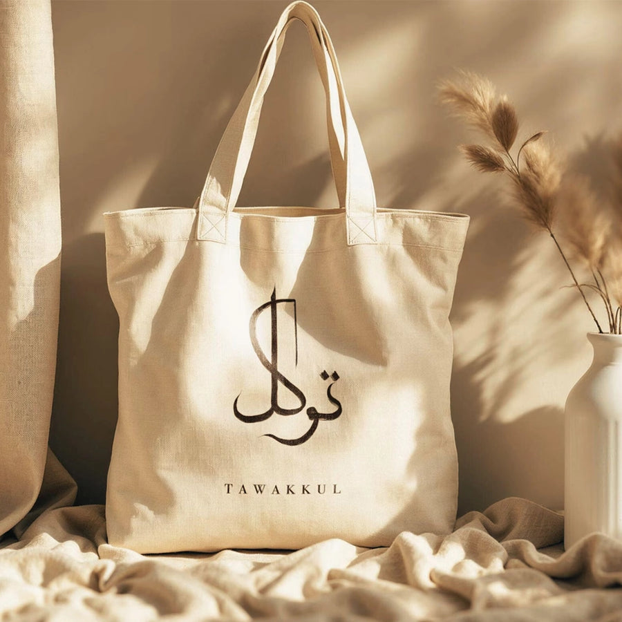 A beige custom tote bag with Arabic script printed on the front, displayed in a sunlit environment with a white bottle and decorative plants in the background.
