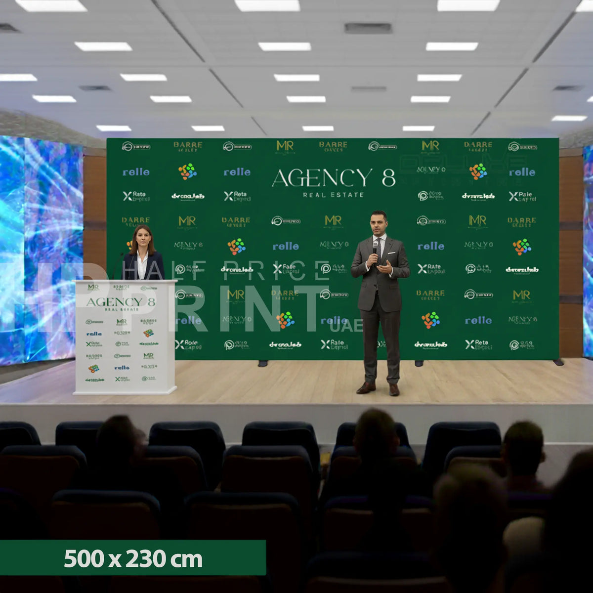 Two speakers on a stage with a green backdrop featuring multiple logos, in an indoor setting.