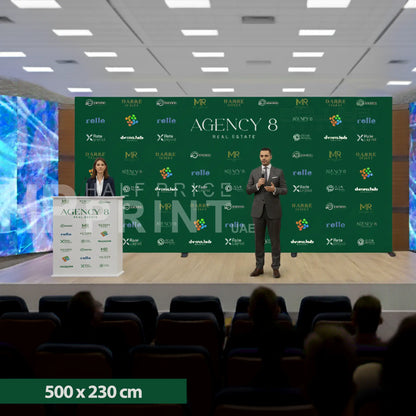 Two speakers on a stage with a green backdrop featuring multiple logos, in an indoor setting.