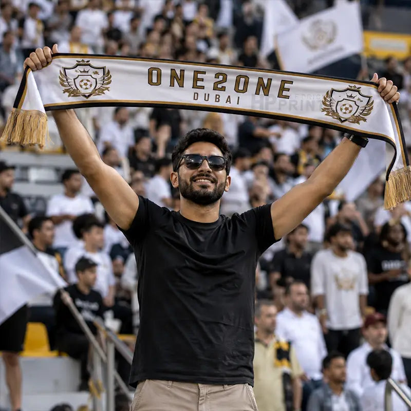 A man in a black t-shirt and sunglasses holds up a white and black 'ONEZONE DUBAI' scarf with gold lettering and tassels in a crowded stadium setting.