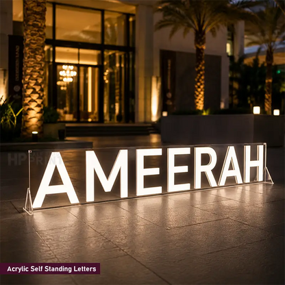 A large acrylic self-standing illuminated sign displaying the name 'AMEERAH' in bright white letters, set outdoors on a tiled floor in front of a modern building at night.