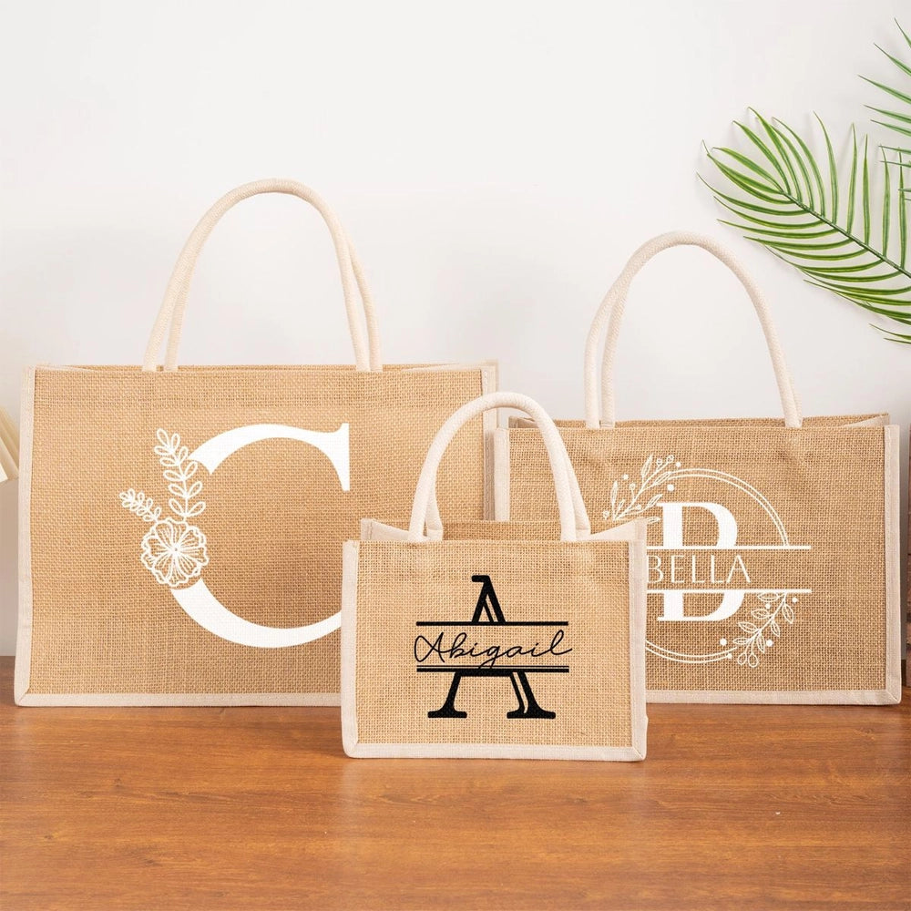 Three natural jute tote bags displayed on a wooden surface, each personalized with a monogram or name in white or black lettering, featuring floral accents and sturdy rope handles.