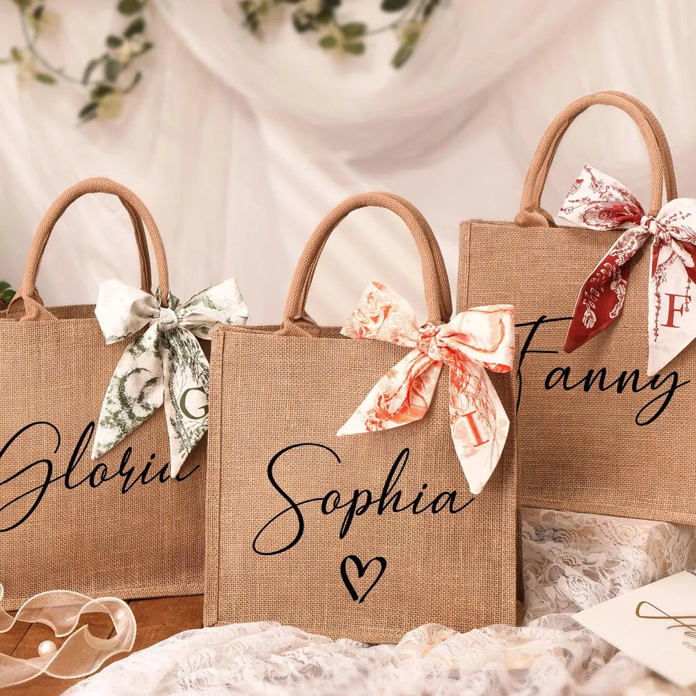Personalized burlap gift bags arranged on a soft fabric backdrop, each printed with a name in elegant script and decorated with patterned ribbon bows tied to the handles.