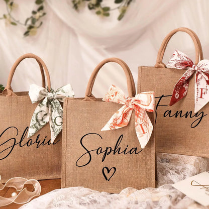 Personalized burlap gift bags arranged on a soft fabric backdrop, each printed with a name in elegant script and decorated with patterned ribbon bows tied to the handles.