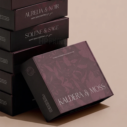 A luxury product packaging display featuring stacked and leaning dark plum-colored boxes with elegant white typography. The front box prominently displays the brand name 'KALDERA & MOSS' alongside the scent 'KALDERA & MOSS SCENTS BY EEA'.