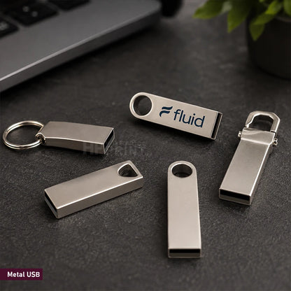 Four silver metal USB flash drives with keychain rings and a cap, arranged on a dark textured surface with a laptop and plant in the background. One drive is printed with the brand name 'fluid'.