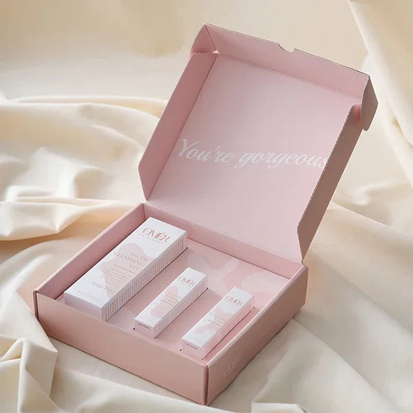 Pink mailer box with inner text 'You're gorgeous' displaying cosmetic packaging on fabric background
