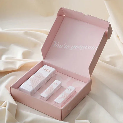 Pink mailer box with inner text 'You're gorgeous' displaying cosmetic packaging on fabric background