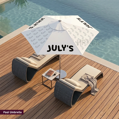 A white pool umbrella with black text reading 'JULY'S' and small line patterns, shown on a wooden deck beside a swimming pool with two lounge chairs and a small side table.