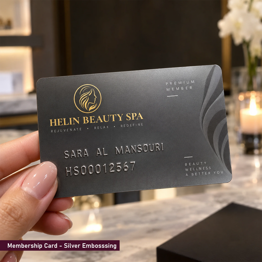 A hand holds a black Helin Beauty Spa premium member membership card with silver embossed text and graphics, set against a softly blurred warm indoor background with a candle and flowers.