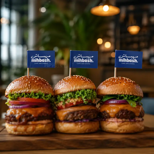 Three gourmet cheeseburgers on a wooden board, each topped with a small blue toothpick flag printed with the brand name 'mibash.' in white, set in a warm, softly blurred restaurant environment.