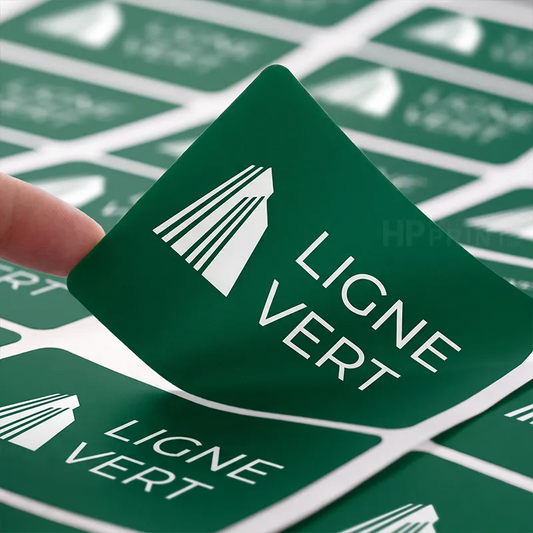 A close-up of a green square sticker being peeled from a sheet, showing the white text 'LIGNE VERT' and a white leaf-like logo. A finger is visible on the left, and more identical stickers are arranged in the background.