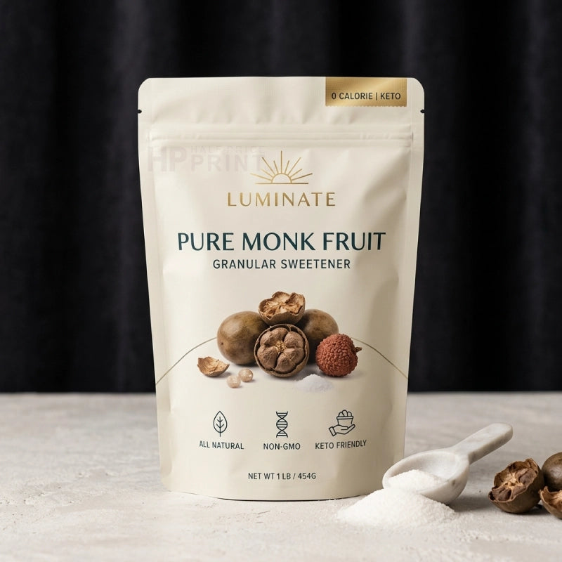 A sealed mylar bag of Luminate Pure Monk Fruit Granular Sweetener on a light stone surface with a dark draped backdrop. The package is off-white with gold and dark blue text, showing fruit illustrations and a small scoop of white powder with monk fruit pieces beside it.