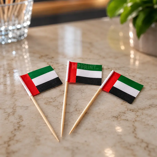 Three small UAE flag toothpick cake toppers on wooden sticks, arranged on a beige marble surface with a blurred glass and green plant in the background.