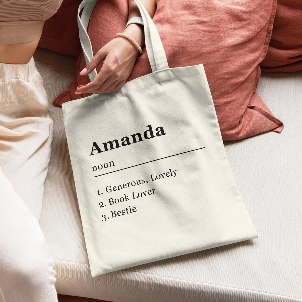 Personalized cream canvas tote bag with the name “Amanda” printed in bold black text, styled like a dictionary definition with descriptive traits, resting on a light sofa with soft pillows.