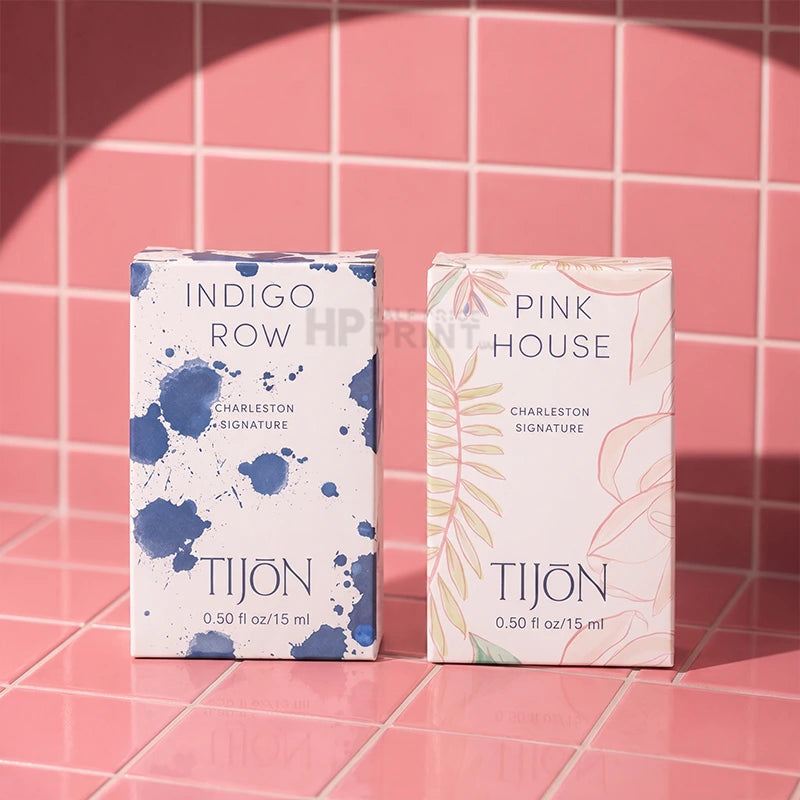 Two Tijon Charleston Signature fragrance tuck top boxes standing upright on a pink tiled surface. The left box is white with blue paint splatter graphics and reads 'INDIGO ROW.