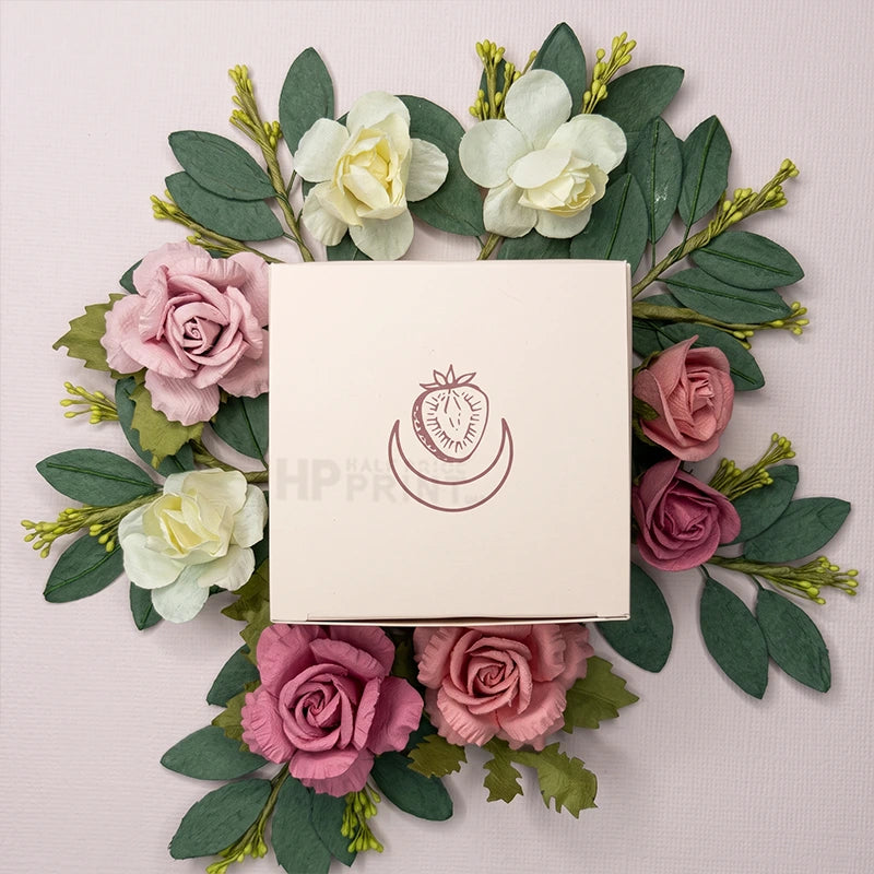 A square cream colored product box with a strawberry graphic logo, surrounded by an arrangement of pink and white artificial flowers and green leaves on a pale pink background.