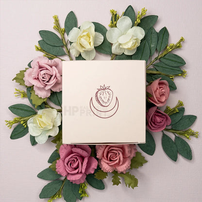 A square cream colored product box with a strawberry graphic logo, surrounded by an arrangement of pink and white artificial flowers and green leaves on a pale pink background.