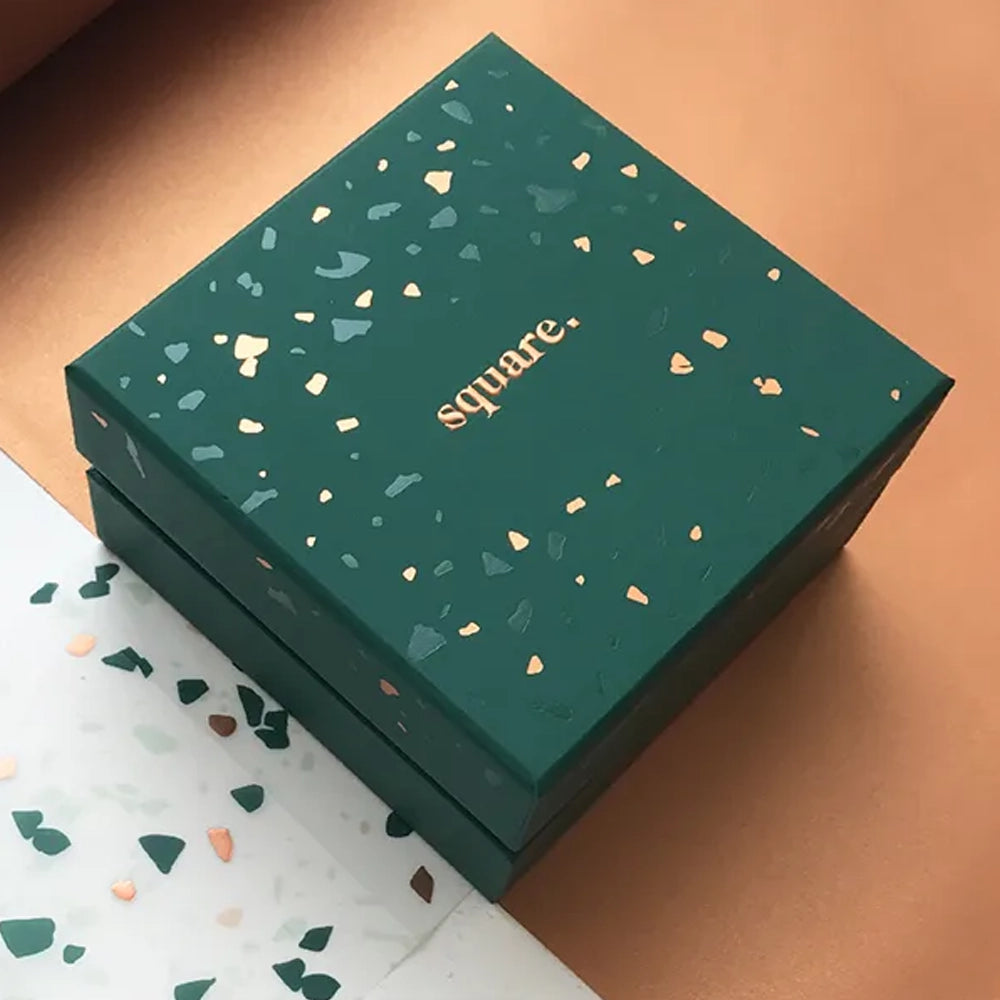 A dark green square gift box with a matching lid, decorated with small speckled accents in light and gold tones, featuring the word “square.” centered on top, placed on a warm neutral background.