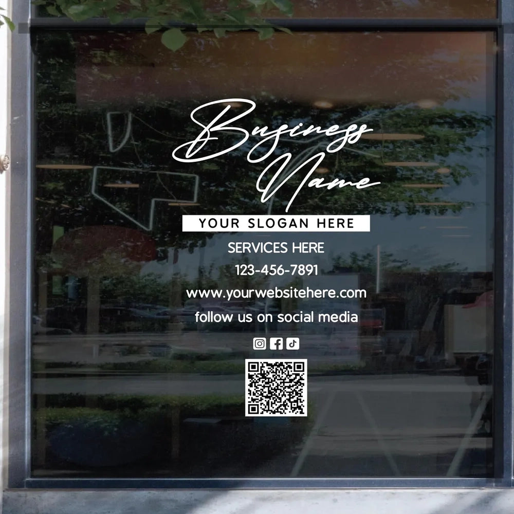 Glass storefront window featuring a business name vinyl sticker with slogan, services text, phone number, website, social media icons, and a QR code, shown from outside with street reflections.